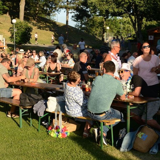 Eine Gruppe von Menschen sitzt an Tischen in einem Außenbereich, möglicherweise einem Park, mit Bäumen und einem Hügel im Hintergrund. Einige tragen Sonnenbrillen und Hüte, und es gibt Getränke und Taschen auf den Tischen.