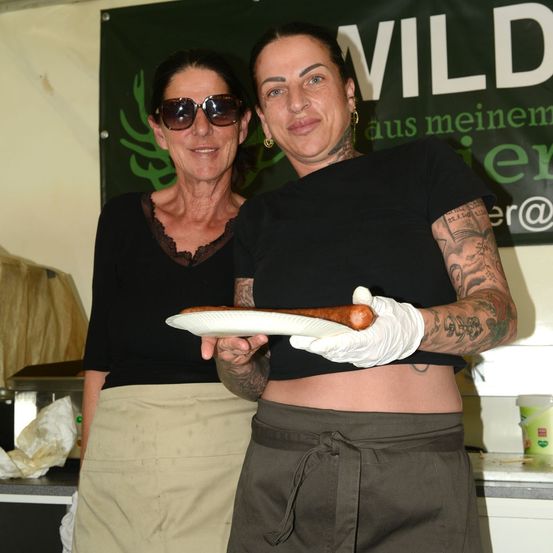 Zwei Frauen in einer Küche, eine hält einen Teller mit einer langen Wurst, die andere lächelt und trägt eine Sonnenbrille. Ein Banner mit dem Wort 'Wild' ist hinter ihnen.
