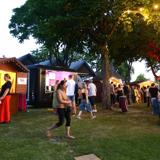 Ein geschäftiges Outdoor-Event mit Food Trucks und Menschen, die sich unter Bäumen bei Dämmerung mischen.