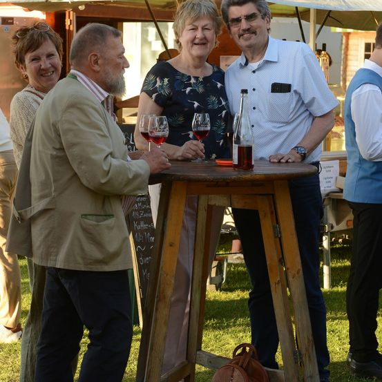 Eine Gruppe von Menschen steht um einen Tisch mit Weinglas und einer Flasche Wein darauf. Sie lächeln und scheinen sich zu amüsieren.