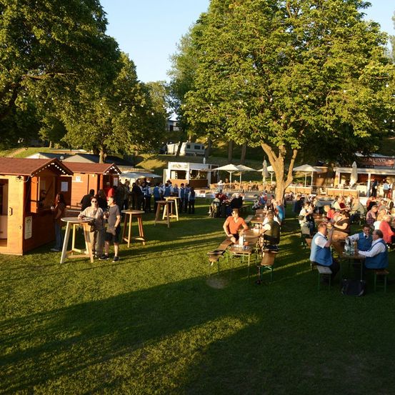 Eine Gruppe von Menschen genießt Getränke und Essen bei einer Outdoor-Veranstaltung im Schatten von Bäumen.