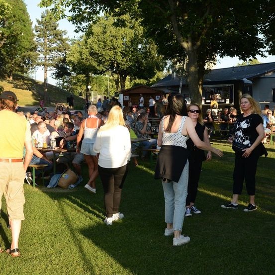 Eine Gruppe von Menschen versammelt sich in einem Park, einige stehen und andere sitzen auf Bänken. Im Hintergrund sind Bäume und ein Gebäude zu sehen.