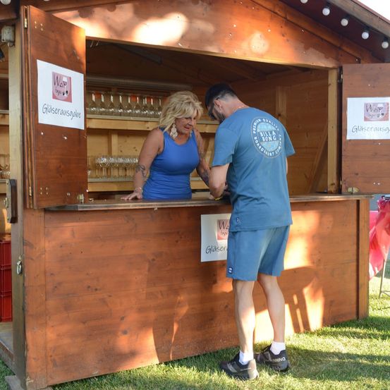 Eine Frau in einem blauen Tanktop steht hinter einem hölzernen Tresen und schaut einem Mann in einem blauen T-Shirt und Shorts an. Sie befinden sich an einem Weinverkostungsstand mit ausgestellten Weingläsern.