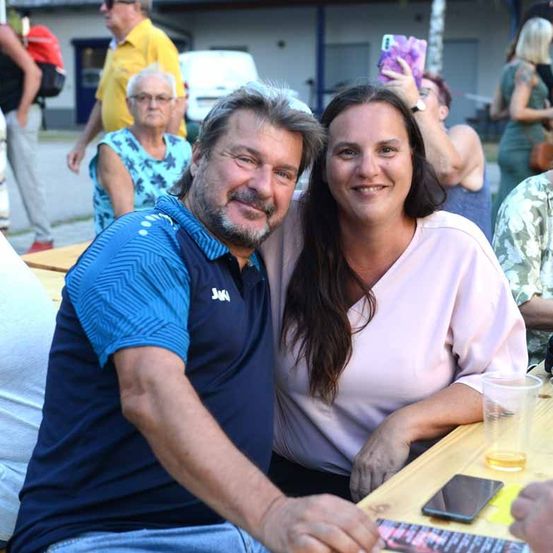 Ein Mann und eine Frau lächeln und posieren für ein Foto bei einer Outdoor-Veranstaltung, mit anderen Leuten und einem Gebäude im Hintergrund.