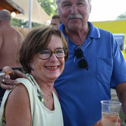 Ein älterer Mann und eine Frau lächeln für ein Foto bei einer Outdoor-Veranstaltung, wobei der Mann ein Getränk hält und die Frau eine Brille trägt.
