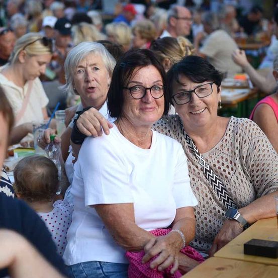 Eine Gruppe von Menschen, darunter zwei ältere Frauen, die lächelnd für ein Foto posieren, sitzen an einem Tisch in einem überfüllten Außenbereich.
