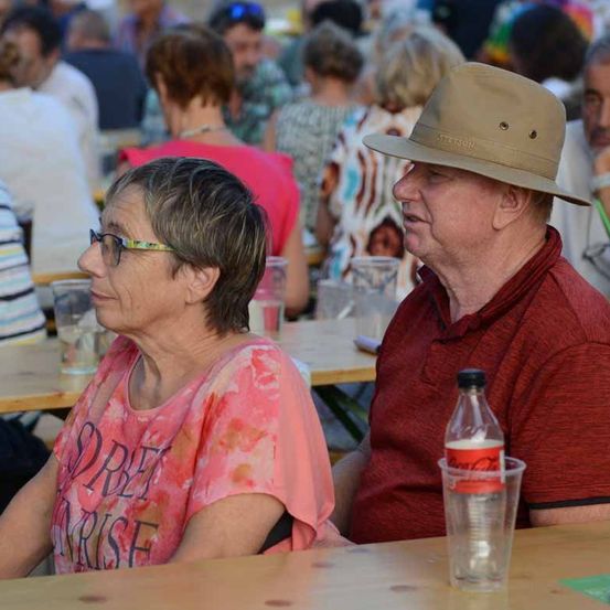 Ein älteres Paar sitzt an einem Tisch in einem Outdoor-Essbereich, der Mann trägt einen Hut und die Frau eine Brille. Hinter ihnen sitzen andere Leute an Tischen, und eine Coca-Cola-Flasche steht auf dem Tisch.
