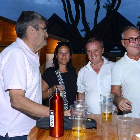 Vier Personen stehen um einen Tisch mit Getränken, zwei Männer in weißen Hemden und zwei Frauen, eine in Schwarz und die andere mit einem Glas.