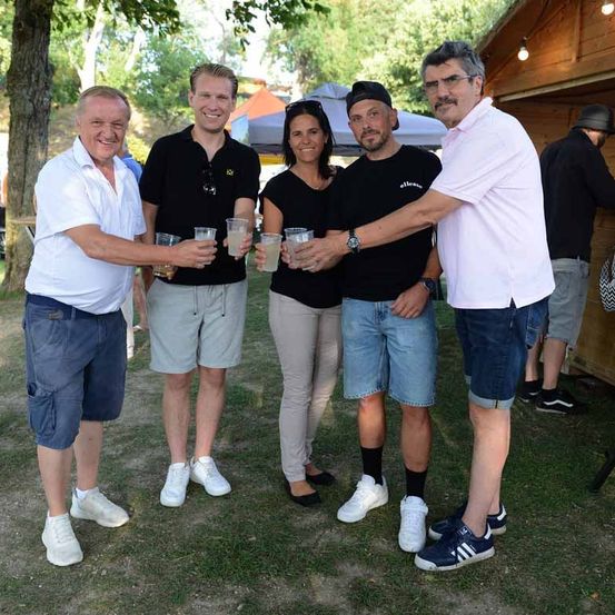 Fünf Menschen stehen auf einer Rasenfläche und halten Getränke in den Händen. Sie lächeln und scheinen für ein Foto zu posieren. Hinter ihnen befindet sich eine Holzstruktur mit Lichtern.