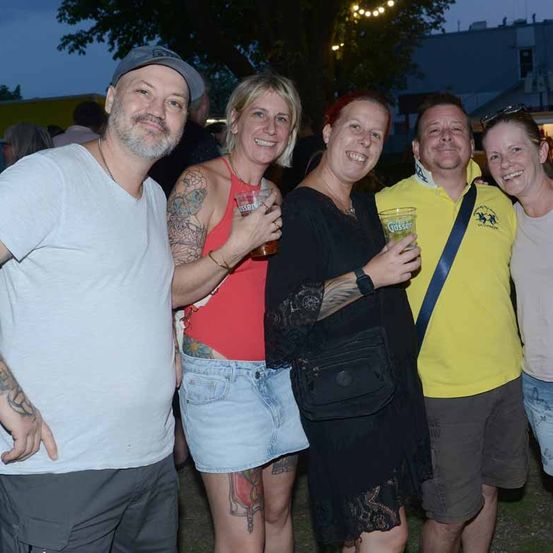 Fünf Erwachsene, vier mit Gläsern in der Hand, lächeln und posieren für ein Foto bei einer Outdoor-Veranstaltung bei Nacht.