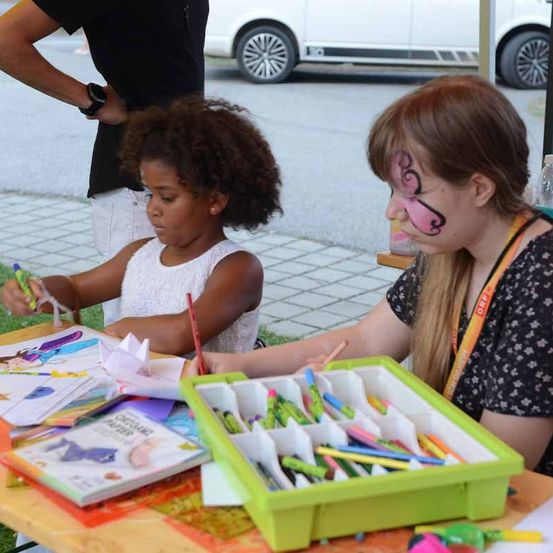 Ein junges Mädchen mit Gesichtsbemalung sitzt mit einer Frau an einem Tisch und beide färben ein Buch mit Buntstiften ein.