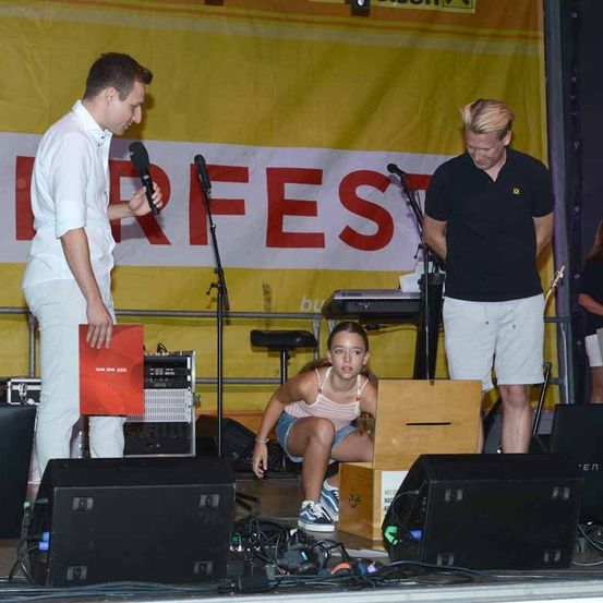 Ein Mann hält eine rote Mappe und spricht in ein Mikrofon auf der Bühne. Ein junges Mädchen sitzt in einer Holzkiste, und ein weiterer Mann steht hinter einem Keyboard. Ein gelber Banner mit roten Buchstaben steht für 'FEST'.