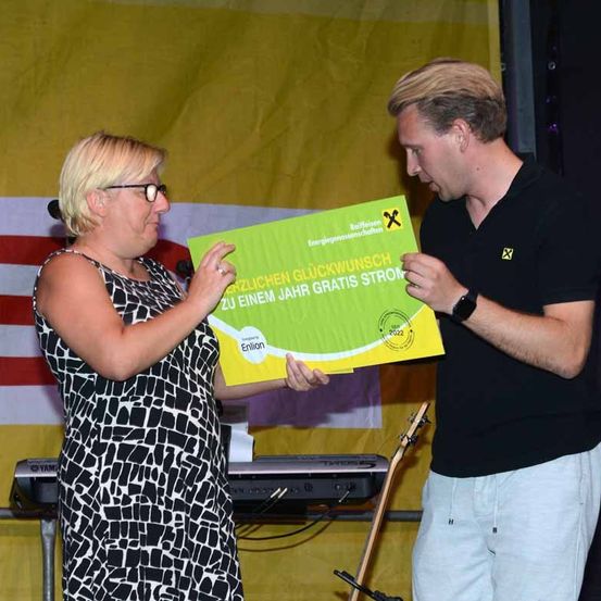 Eine Frau in einem schwarz-weißen Kleid und ein Mann in einem schwarzen Hemd halten ein grünes Zertifikat mit dem Text 'Herzlichen Glückwunsch zu einem Jahr gratis Strom'. Sie stehen vor einem gelben Hintergrund mit einer rot-weißen Flagge.