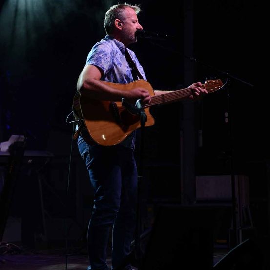 Ein Mann auf der Bühne spielt eine akustische Gitarre und singt in ein Mikrofon unter Bühnenlicht.