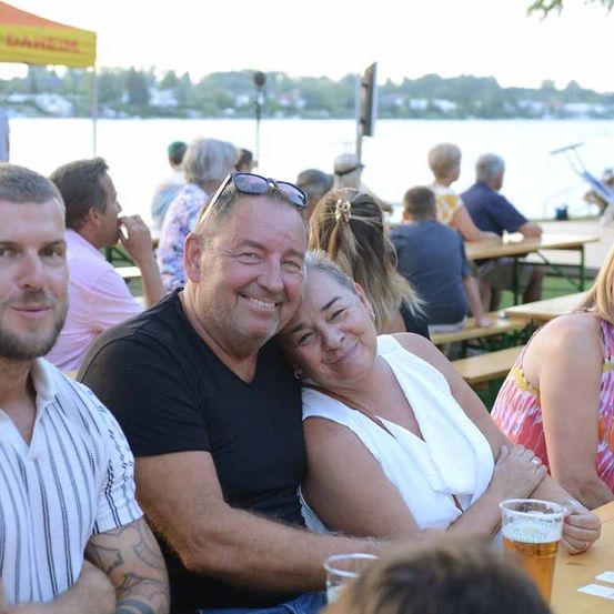 Eine Gruppe von Menschen sitzt an einem Picknicktisch am See und lächelt für ein Foto. Der Mann hat eine Sonnenbrille auf dem Kopf.