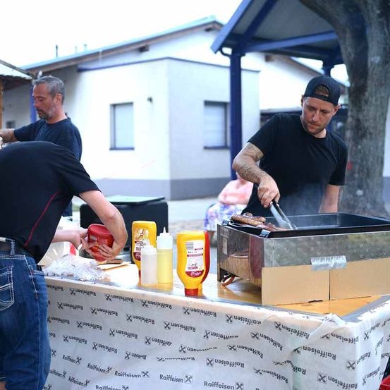 Drei Männer grillen Essen auf einem Tisch mit Gewürzen und einem Gebäude im Hintergrund.