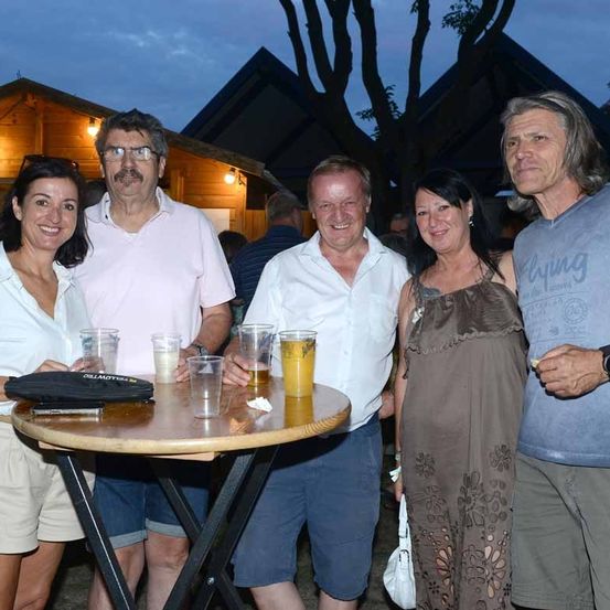 Eine Gruppe von Menschen steht um einen Tisch mit Getränken, lächelt und posiert für ein Foto bei einer Veranstaltung im Freien.