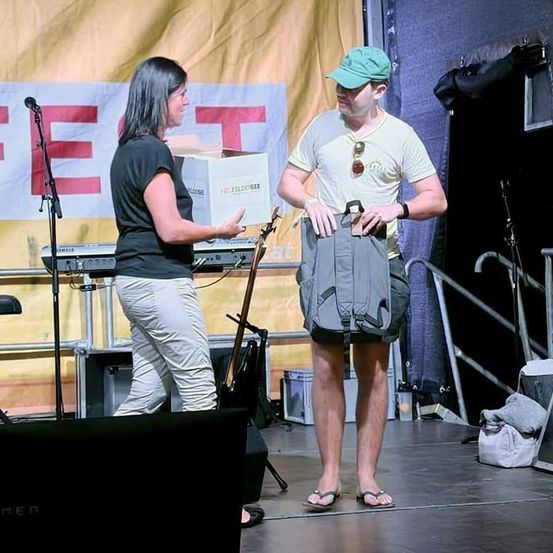 Eine Frau übergibt einem Mann auf der Bühne eine Box, mit einem Banner im Hintergrund, das 'FEST' steht.