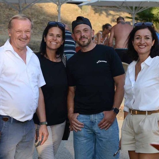 Vier Personen stehen zusammen, lächeln und posieren für ein Foto. Der Mann auf der linken Seite trägt ein weißes Hemd, und die Frau neben ihm trägt ein schwarzes Oberteil. Der Mann in der Mitte trägt ein schwarzes T-Shirt mit dem Wort 'ellesse' darauf. Die Frau auf der rechten Seite trägt ein weißes Blusenhemd.