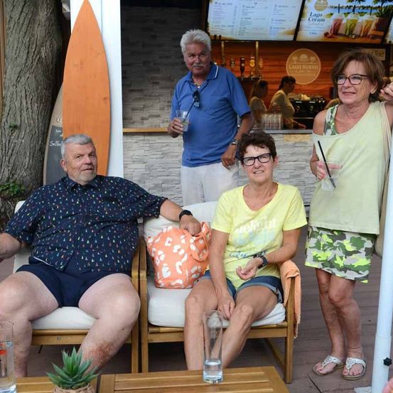 Vier Menschen sitzen in Stühlen vor einer Bartheke, zwei Männer und zwei Frauen. Der Mann links trägt ein Blumenhemd und Shorts, während die Frau rechts ein grünes Kleid trägt.