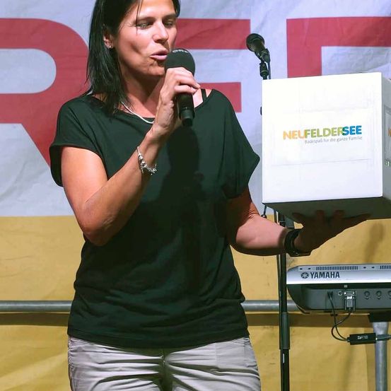Eine Frau in einem schwarzen Shirt spricht in ein Mikrofon auf einer Bühne mit einem Yamaha-Keyboard und einer Box mit der Aufschrift 'Neufeldersee' hinter ihr.