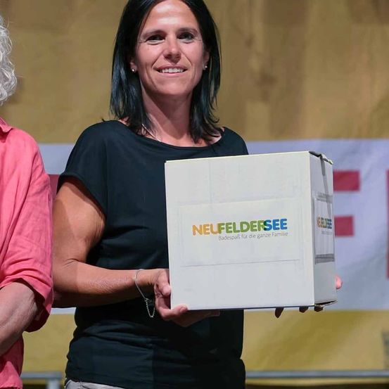 Eine Frau hält eine Box mit der Aufschrift 'NeuFeldersee' und lächelt, neben einer weiteren Person in einem rosa Hemd.