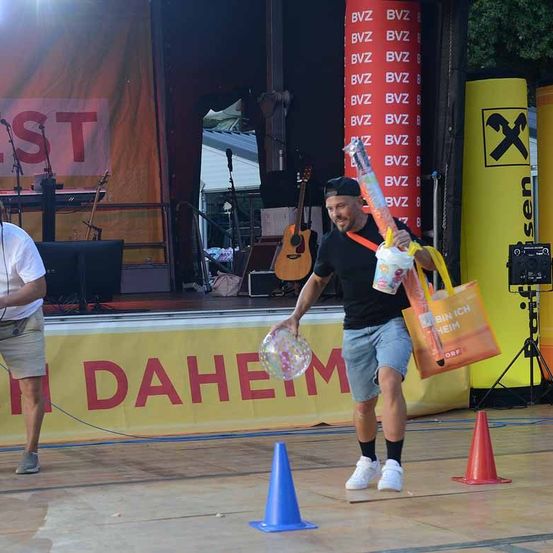 Ein Mann auf der Bühne hält einen Ballon und eine Tasche, mit einem Banner, das 'Daheim' dahinter steht, und eine Person steht links.