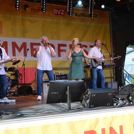 Eine Band tritt auf der Bühne mit einem gelben Hintergrund mit roten Buchstaben auf. Die Sängerin ist eine Frau in einem grünen Kleid, während die anderen Bandmitglieder Männer sind, die Gitarre und Keyboard spielen.