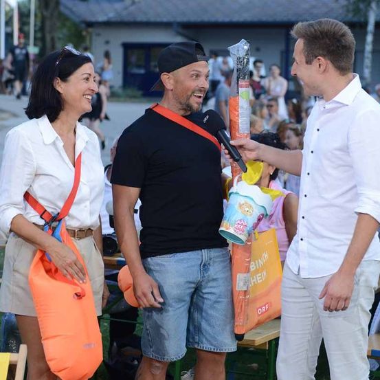 Ein Mann in einem schwarzen T-Shirt und Jeansshorts hält ein Mikrofon und eine Tasche, während ein anderer Mann in einem weißen Hemd einen Plastikbeutel hält. Eine Frau in einem weißen Blusenkleid und einer roten Tasche steht in der Nähe. Sie alle lächeln und scheinen sich bei einer Veranstaltung im Freien zu befinden.
