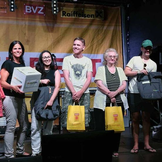 Fünf Personen stehen lächelnd auf einer Bühne, eine hält eine Box und die anderen tragen Taschen. Ein Banner mit den Texten 'Raiffeisen' und 'BVZ' ist hinter ihnen.