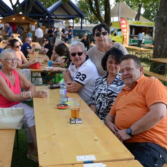 Eine Gruppe von Menschen sitzt an einem Tisch in einem Restaurant im Freien, einige mit Sonnenbrillen und lächelnd. Es gibt Getränke und Essen auf dem Tisch, und ein Imbisswagen ist im Hintergrund.