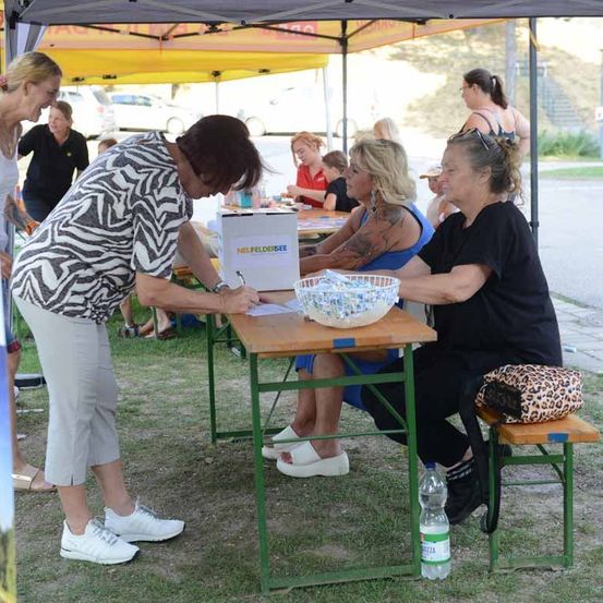 Eine Frau unterzeichnet eine Petition an einem Tisch unter einem Zelt, während andere Leute sitzen und stehen.