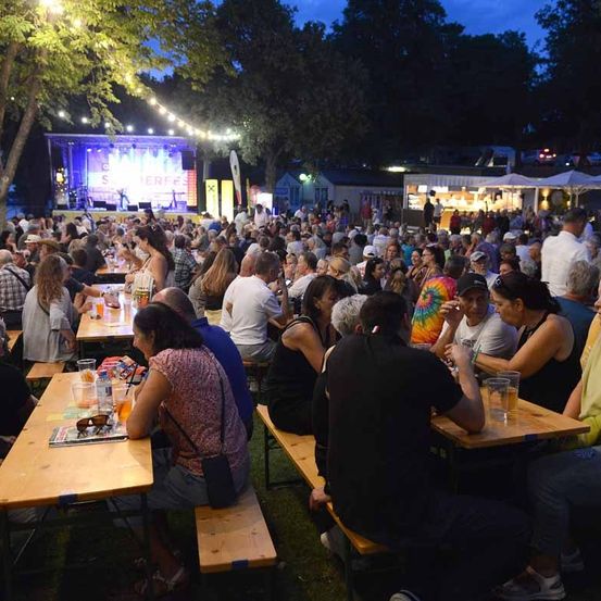 Ein nächtliches Outdoor-Festival mit Bühne, Menschen an Tischen und Imbissständen im Hintergrund.