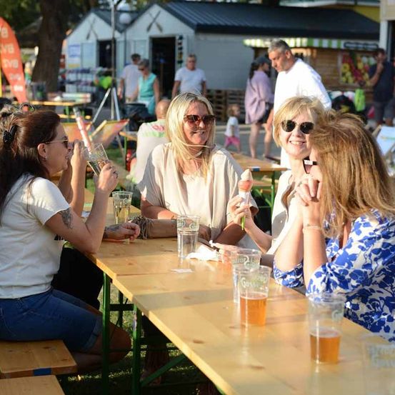 Vier Frauen sitzen an einem Picknicktisch in einem Park, trinken Bier und essen Eis. Der Tisch hat Gläser und Tassen. Im Hintergrund gehen Menschen und ein Gebäude mit einem Schild.