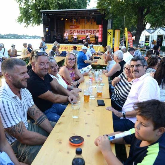 Eine Gruppe von Menschen sitzt an Tischen in einem Außenbereich, möglicherweise bei einem Festival. Sie lächeln und scheinen sich zu amüsieren. Auf den Tischen stehen Gläser, Flaschen und ein Handy. Im Hintergrund befindet sich eine Bühne mit einem gelben Banner und einer Person, die steht.