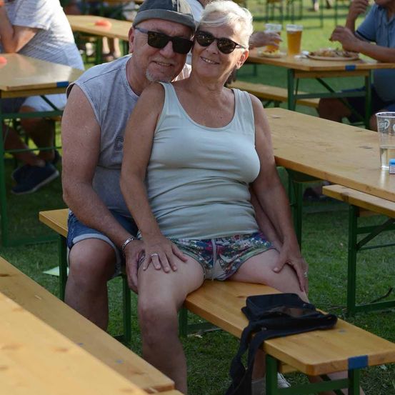 Ein älteres Paar sitzt zusammen auf einer Bank in einem Biergarten im Freien, beide tragen Sonnenbrillen und lächeln für die Kamera.