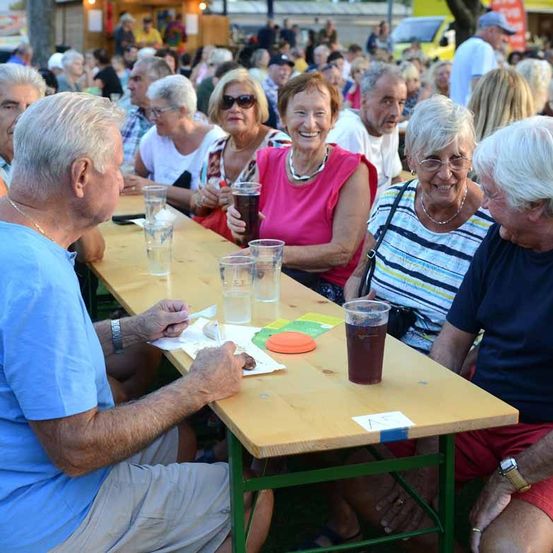 Eine Gruppe älterer Menschen sitzt an einem langen Tisch, wahrscheinlich bei einem Festival, genießt Getränke und Essen. Sie lächeln und unterhalten sich miteinander.