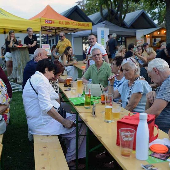 Eine Gruppe von Menschen sitzt an einem langen Tisch bei einer Outdoor-Veranstaltung, trinkt Bier und unterhält sich. Die Umgebung umfasst Zelte und einen Rasenbereich.