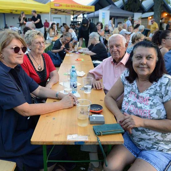 Eine Gruppe von Menschen sitzt an einem langen Tisch im Freien, lächelt und posiert für ein Foto. Auf dem Tisch stehen Wasserflaschen, Gläser und eine Geldbörse. Im Hintergrund befinden sich Zelte und Menschen, die auf Bänken sitzen.
