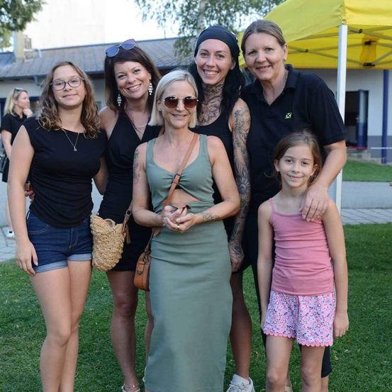 Eine Gruppe von Frauen und ein junges Mädchen stehen lächelnd auf einem Rasenfeld und posieren für ein Foto. Sie sind lässig gekleidet, und ein gelbes Zelt ist im Hintergrund zu sehen.
