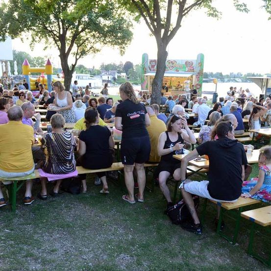 Eine große Gruppe von Menschen sitzt an Picknicktischen in einem Park und genießt Essen und Getränke. Im Hintergrund befinden sich Bäume, ein Hüpfburg und ein Imbissstand.