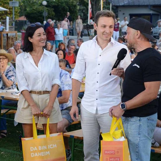 Ein Mann und eine Frau mit Einkaufstüten werden von einem Mann mit Mikrofon interviewt. Sie stehen auf einer Wiese mit Tischen und Leuten, die auf Bänken hinter ihnen sitzen.