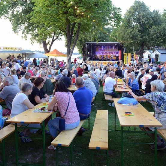 Eine große Menschenmenge sitzt auf Picknicktischen in einem Park, genießt Essen und Getränke und sieht sich eine Band auf der Bühne vor.