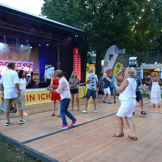 Eine Gruppe von Menschen tanzt auf einer Holzbühne, während eine Band im Hintergrund spielt. Die Bühne ist von Bäumen umgeben und hat ein gelbes Banner mit Text.