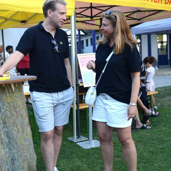 Ein Mann und eine Frau stehen vor einem Baum, beide in schwarzen Shirts und weißen Shorts gekleidet. Der Mann hält einen Tisch, während die Frau eine weiße Handtasche trägt. Hinter ihnen befindet sich ein gelbes Zelt und ein Gebäude.