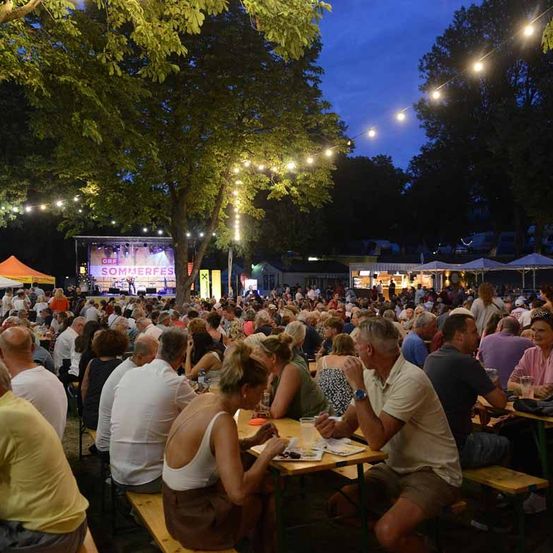 Eine nächtliche Freiluftveranstaltung mit Bühne und Lichterketten. Die Menschen sitzen an Tischen und Bänken und genießen Essen und Getränke. Die Bühne hat eine große Leinwand mit der Aufschrift 'SOMMERFEST'.