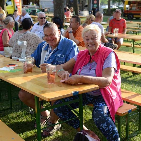 Ein älteres Paar sitzt an einem Holztisch in einem Biergarten im Freien, lächelt und posiert für ein Foto. Sie halten Gläser mit Bier in den Händen. Hinter ihnen sitzen andere Menschen an Tischen, einige mit Essen und Getränken. Die Umgebung ist im Freien mit Bäumen und Gras.