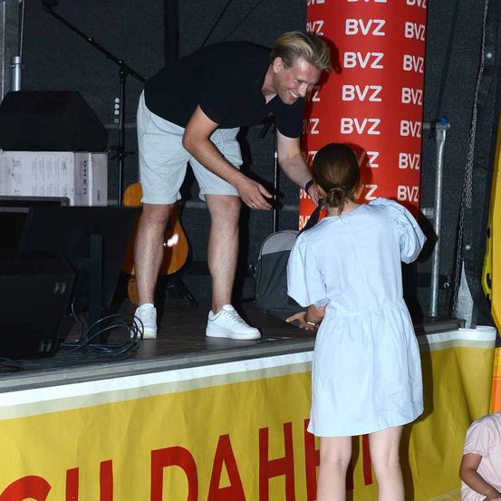Ein Mann auf der Bühne beugt sich vor, um mit einer Frau in einem weißen Kleid zu sprechen. Dahinter befindet sich ein roter Banner mit weißem Text und einem gelben Hintergrund.