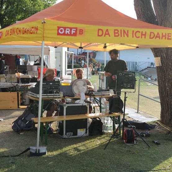 Zwei Männer sitzen unter einem orange-gelben Zelt mit der Aufschrift 'ORF B DA BIN ICH DAHE'. Sie sind von verschiedenen elektronischen Geräten und einem Ventilator umgeben. Ein Mann steht in der Nähe, und im Hintergrund befindet sich ein Gebäude mit einer Treppe.