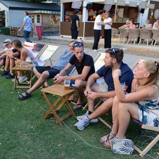 Eine Gruppe von Menschen sitzt auf Liegestühlen auf einem Rasen, möglicherweise bei einem Festival. Einige trinken Bier und einige schauen auf ihre Telefone.